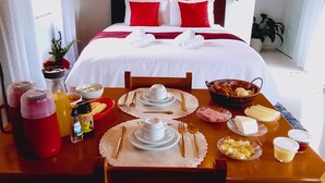 A neatly arranged breakfast table with various food items and utensils, a bed with red and white bedding, and a potted plant in the corner. - Refuge in the mountains/near the trail to Pico do Papagaio Garcias Peak and Waterfall (Aiuruoca)