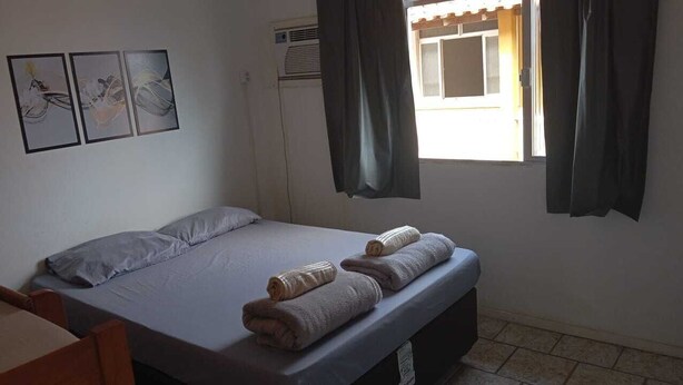Una cama individual con colchón y almohadas grises, dos toallas enrolladas, una mesita y una ventana con cortinas.