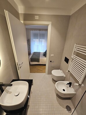 A modern bathroom with a sink, bidet, and toilet. There is a door leading to a bedroom with a bed and a window with curtains. - Lilla Suites - Musei Vaticani (Rome)