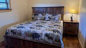 A bed with a quilt featuring animal prints, a wooden headboard, a bedside table with a lamp, and a window with blinds. - Wharton Wilderness Ranch House (South Fork)