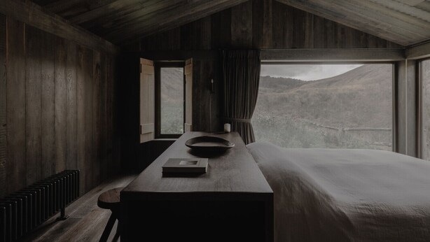 Una habitación de cabaña rústica con interior de madera, una cama grande, una mesita de noche, una ventana con cortinas y vistas a un paisaje montañoso.