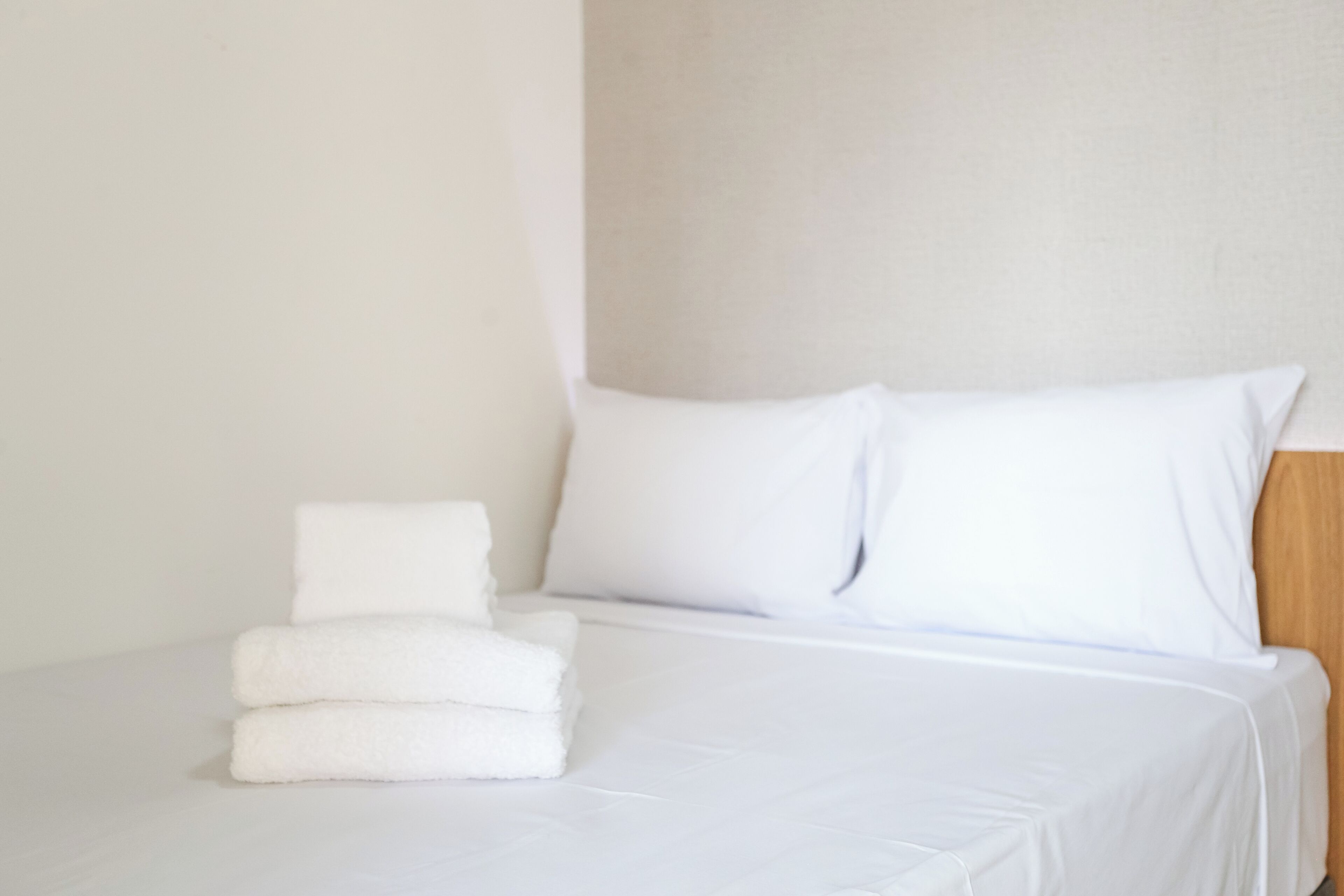 A neatly made bed with white linens and pillows, a stack of folded white towels, and a wooden headboard.