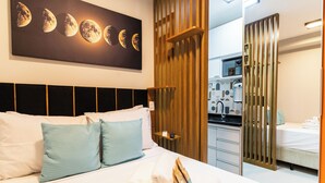 Habitación de hotel con cama, almohadas, un cuadro con motivos lunares, un panel de madera y una vista parcial de una cocina americana.