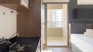 Habitación de hotel moderna con cocina, balcón y vistas a edificios, además de una cama con televisor.