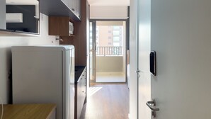 Un pasillo estrecho con un kitchenette a la izquierda, una puerta a la derecha que da a un balcón, y vista de un edificio al exterior.