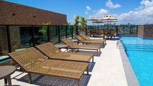 Ein Poolbereich auf dem Dach mit Liegestühlen und Sonnenschirmen, umgeben von Glaswänden mit Blick auf die Stadt.