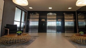 Eine moderne Lobby mit zwei Aufzügen, einem Empfangstresen und zwei runden Tischen mit Pflanzen.