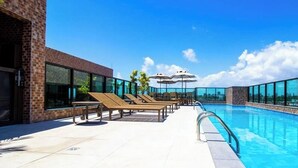 Ein Pool auf dem Dach mit Liegestühlen und Sonnenschirmen, umgeben von großen Fenstern mit Blick auf die Stadt.