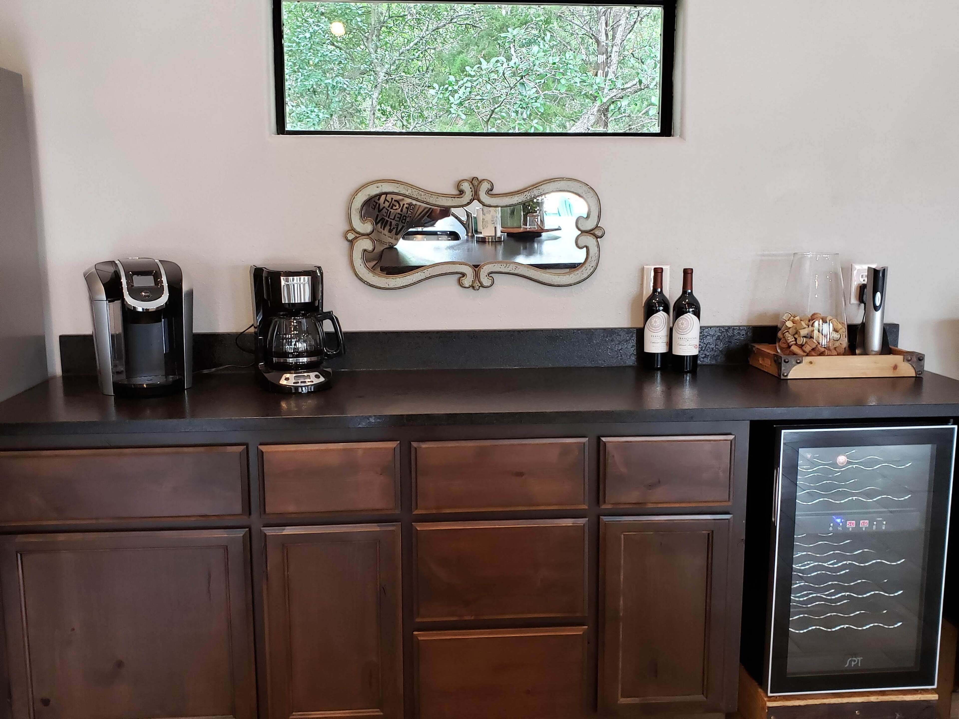A kitchen counter with a coffee maker, two wine bottles, and a decorative mirror.