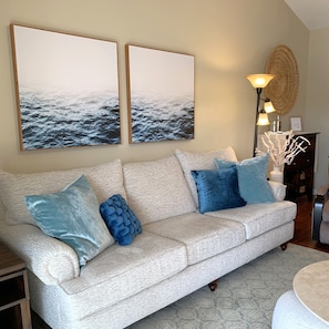 A living room with a beige sofa, blue and white cushions, two framed artworks, a floor lamp, and a woven basket on a wooden table.