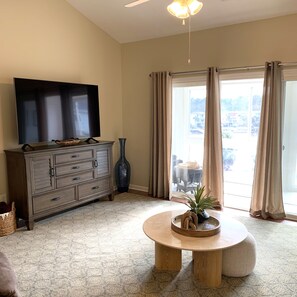 A living room with a flat-screen TV on a wooden cabinet, a round wooden table with a plant, and a view of a balcony with a table and chairs.