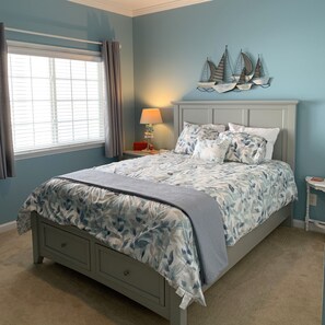 A bedroom with a bed, a nightstand, a lamp, a window with curtains, and a wall decoration.