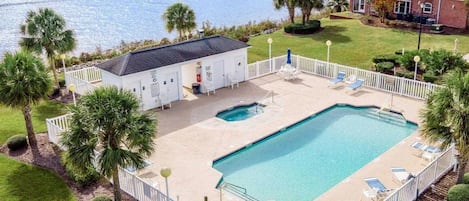 Una piscina rectangular con una terraza blanca, rodeada por una cerca blanca, y una vista a un cuerpo de agua con casas al fondo.