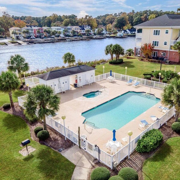 A rectangular swimming pool with a white deck, surrounded by a white fence, and a view of a body of water with houses in the background.
