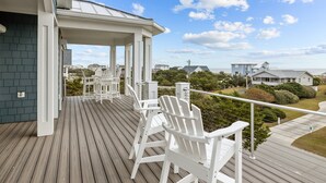 A wooden deck with white Adirondack chairs, a covered area, and a view of the ocean and neighboring houses. - Ocean Views & Surf Fishing Paradise at The Point – Perfect Family Escape! (Emerald Isle)
