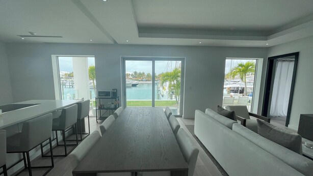 A modern kitchen with a long dining table, bar area, and large windows offering a view of a pool and palm trees. - The Seashell Condo (Nassau)