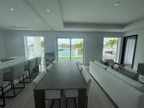 Une cuisine moderne dotée d’une longue table à manger, d’un coin bar et de grandes fenêtres offrant une vue sur une piscine et des palmiers.