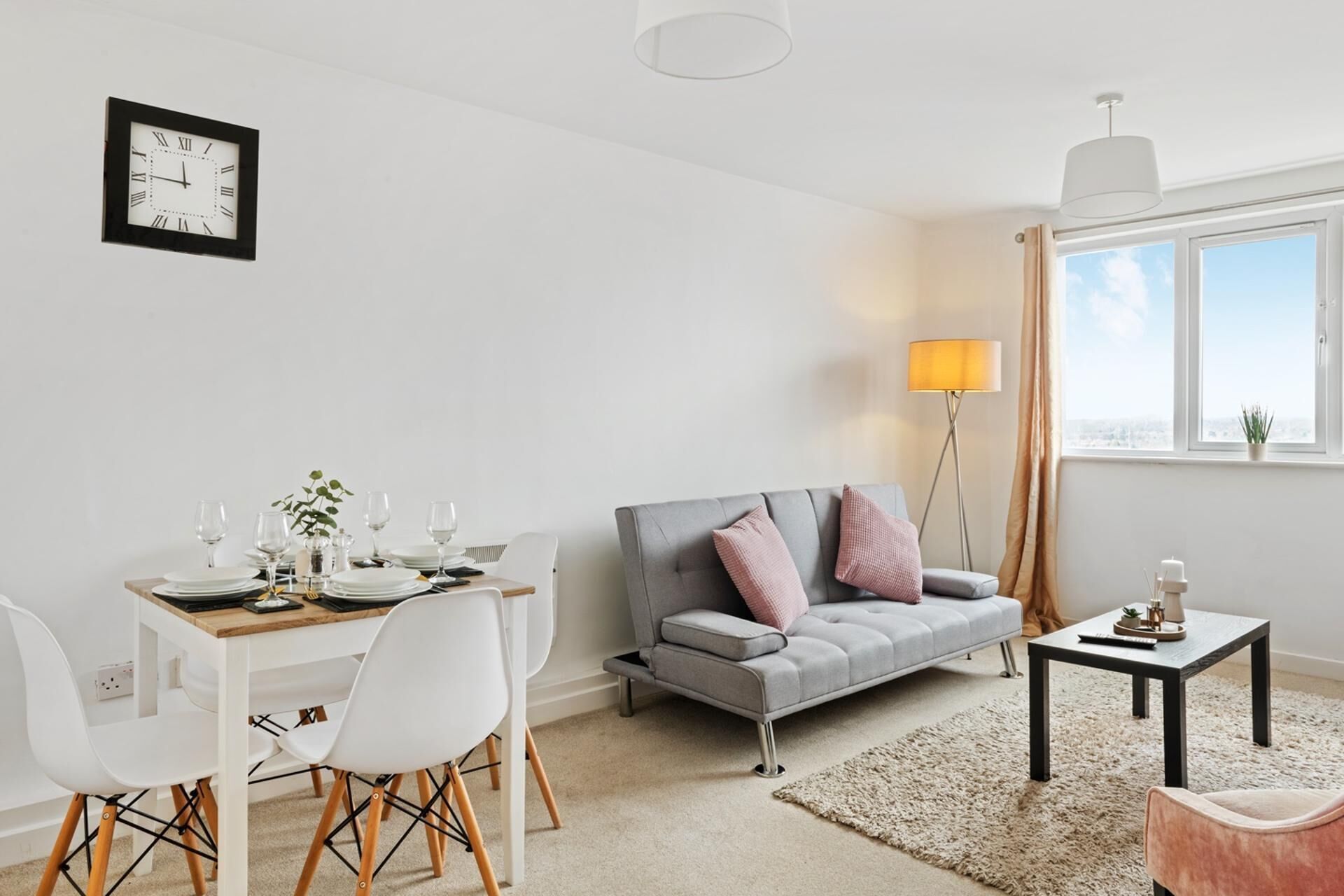 Ein modernes Wohnzimmer mit Essbereich, grauem Sofa, weißen Stühlen und einem kleinen Tisch mit Lampe.