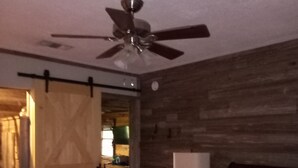 Quarto com cama, ventilador de teto, parede de madeira e porta que dá para uma cozinha.