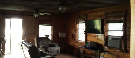 A rustic cabin interior with a wooden ceiling, a sofa, a chair, and a television on a stand.