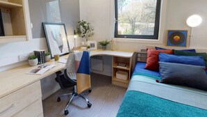 Une chambre moderne avec un bureau, un lit agrémenté de coussins colorés, une fenêtre donnant sur des arbres et une étagère avec des livres et des plantes.