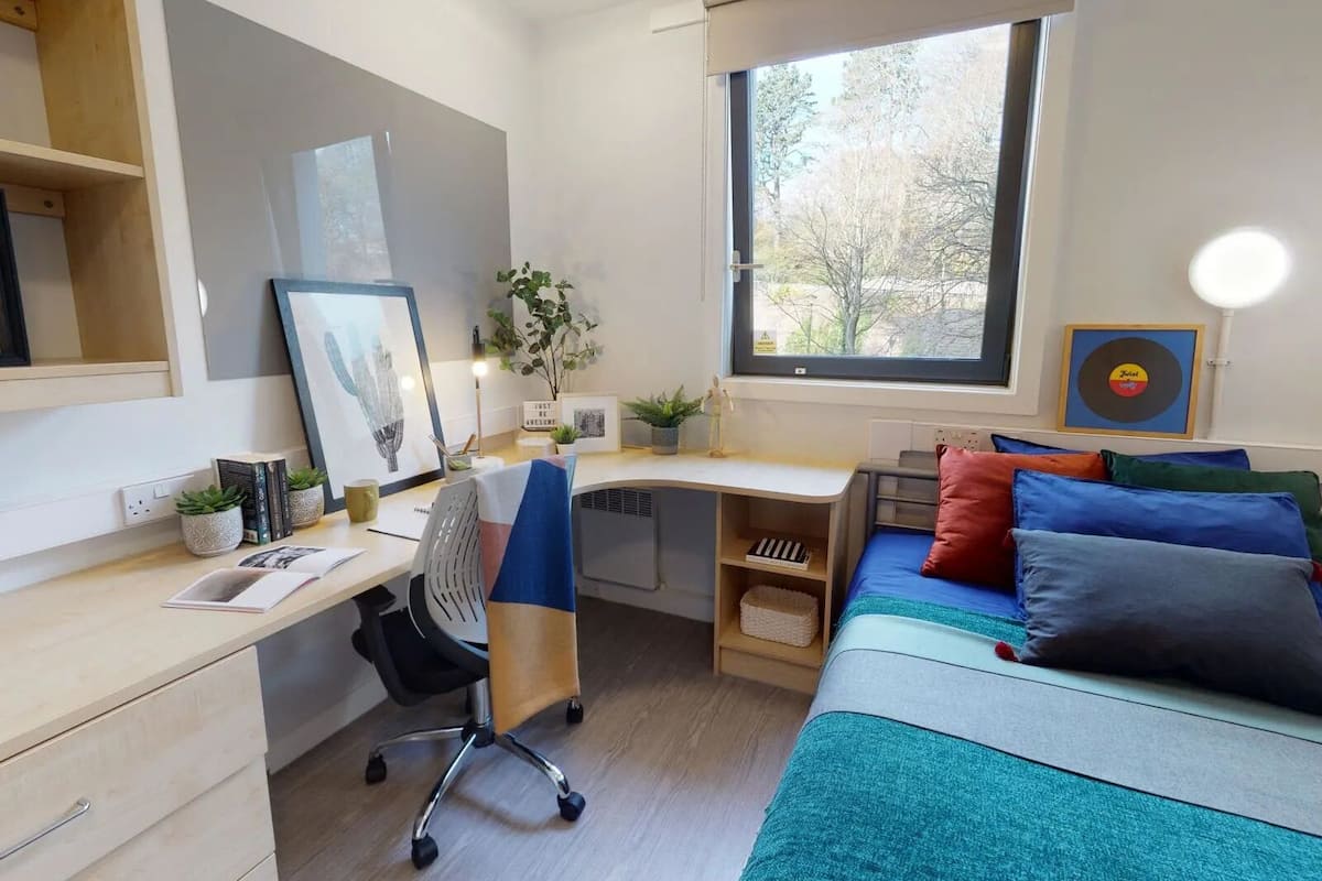 A modern bedroom with a desk, a bed with colorful pillows, a window with a view of trees, and a shelf with books and plants.