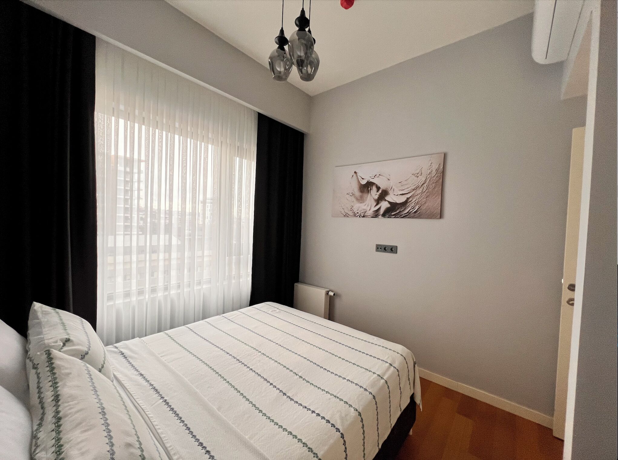 A bedroom with a bed, a window with sheer curtains, a wall-mounted light fixture, and a painting.