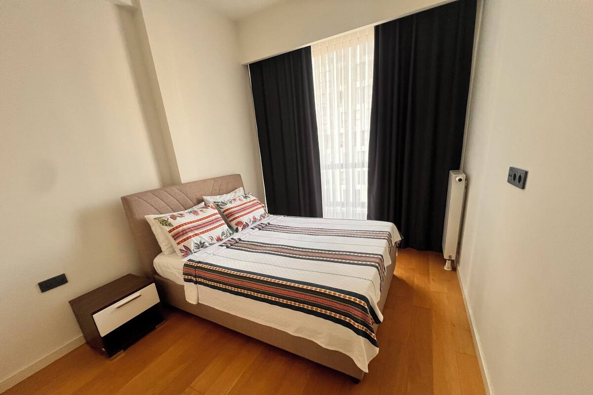 Un lit double avec une couvre-lit à rayures, deux oreillers, une table de chevet et une fenêtre avec des rideaux noirs.