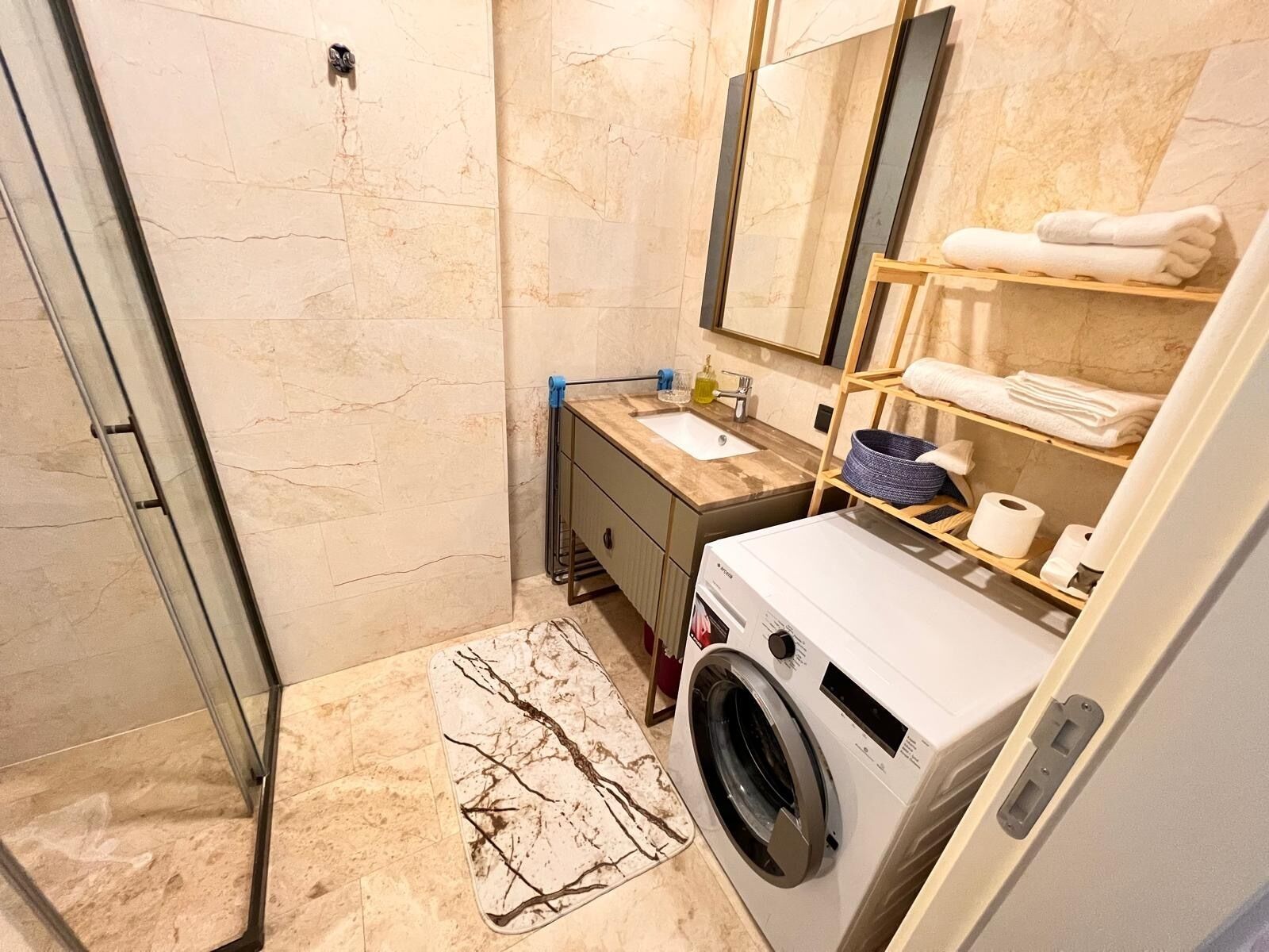 A modern bathroom with a glass shower enclosure, a built-in washing machine, a marble-tiled floor, and a wooden vanity with a mirror.