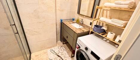 A modern bathroom with a glass shower enclosure, a built-in washing machine, a marble-tiled floor, and a wooden vanity with a mirror.
