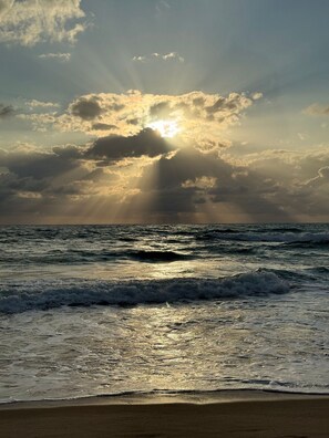 Ein Strand mit Wellen, sandigem Ufer und bewölktem Himmel mit Sonnenschein.