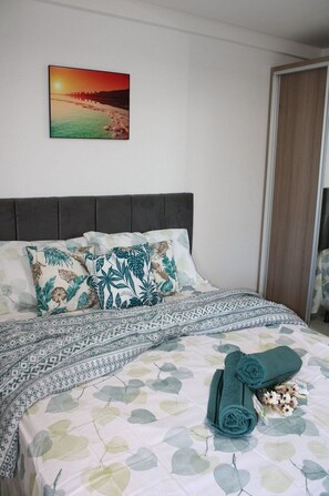 Ein ordentlich bezogenes Bett mit gemusterten Betttextilien, ein Kissen mit tropischem Motiv und ein Sonnenuntergangsbild an der Wand.