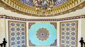Un gran pasillo con intrincados trabajos de azulejos azules y dorados, una lámpara de araña ornamentada y un diseño de medallón central en el techo.