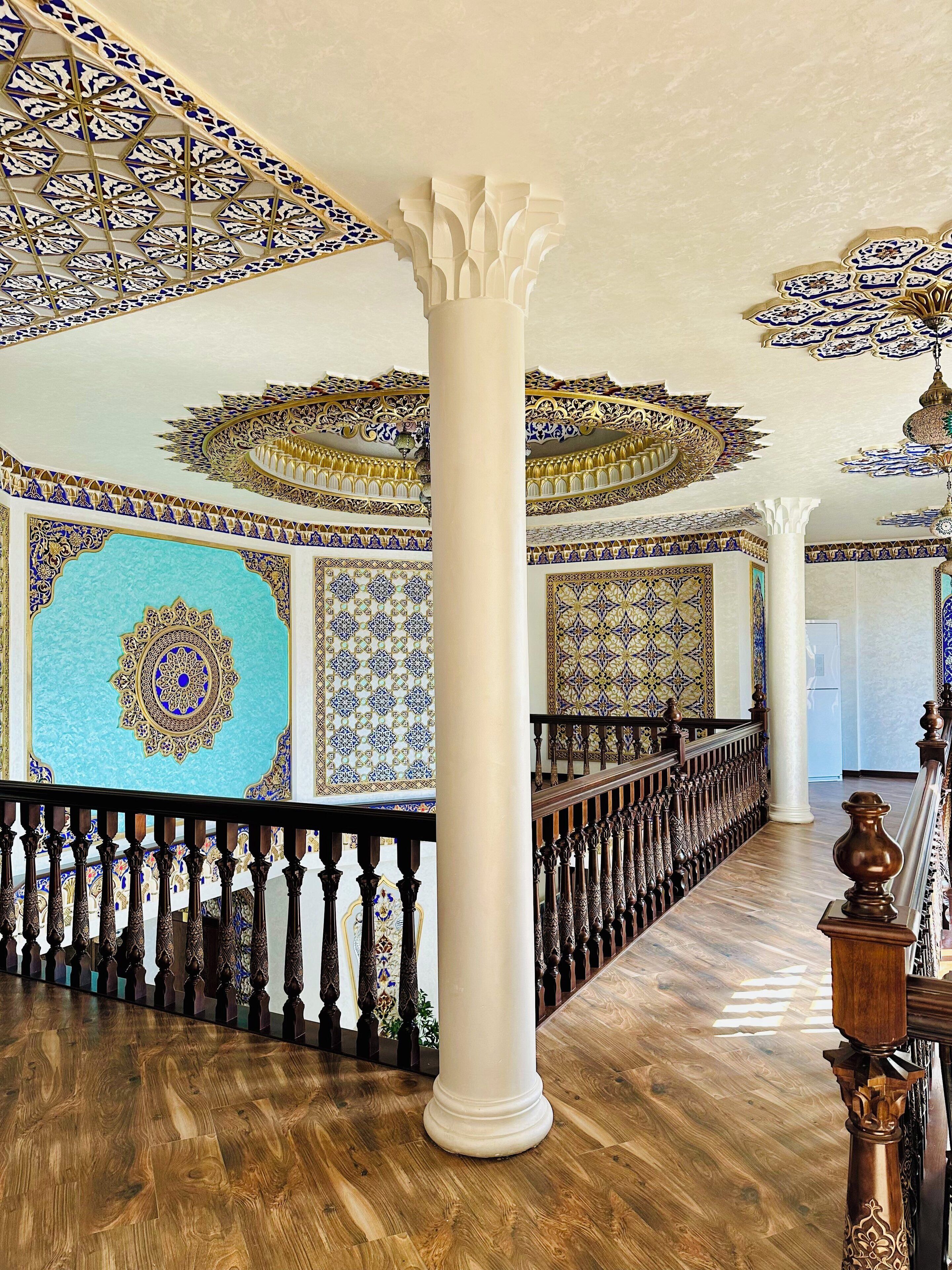 A grand hall with ornate blue and gold patterns on the walls and ceiling, a wooden floor, and a central white column.