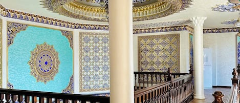 Un salón imponente con elaborados diseños azules y dorados en las paredes y el techo, un suelo de madera y una columna blanca central.
