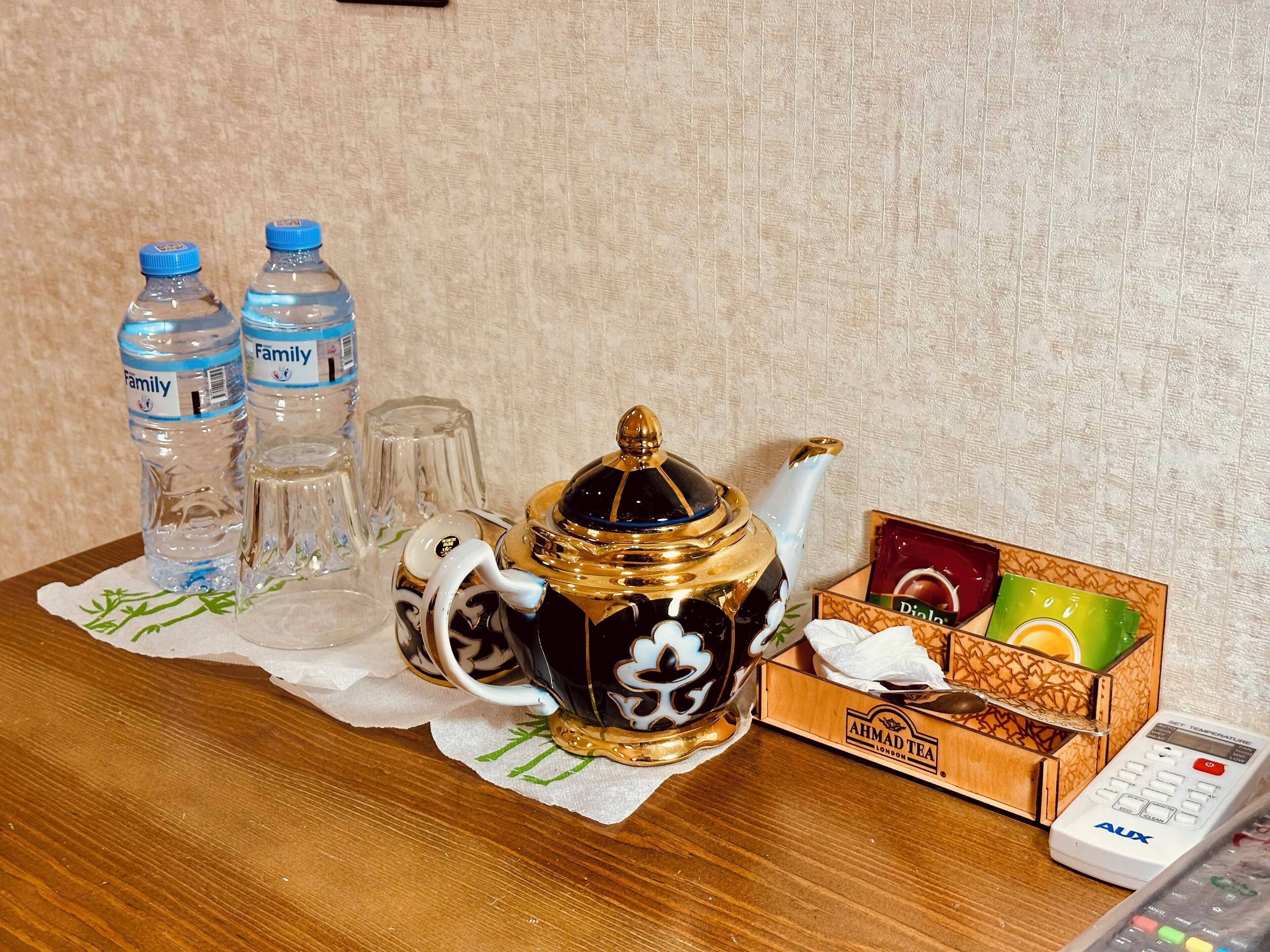 A wooden table with a decorative teapot, two water bottles, a glass, a box of tea, and a remote control.