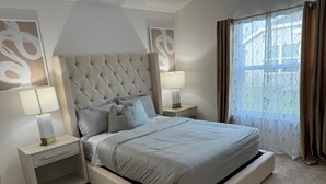 A bedroom with a tufted headboard, two bedside tables with lamps, a bed with a grey bedspread, and a window with curtains.