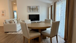 A modern living room with a round wooden dining table, beige upholstered chairs, a flat-screen TV, and a sofa with decorative pillows.