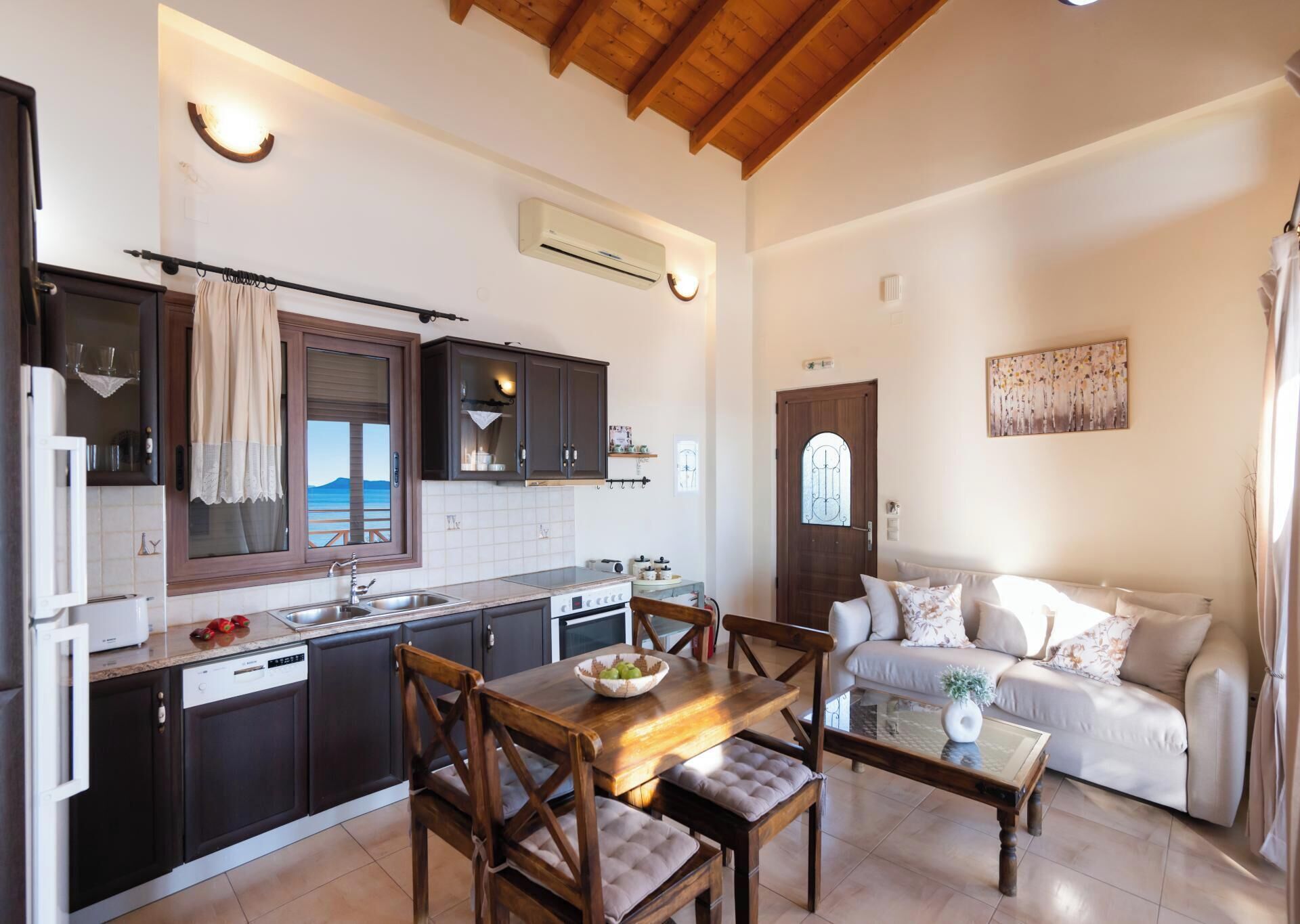 A compact kitchen with dark cabinets, a dining table with chairs, a built-in oven, and a large window overlooking the sea.