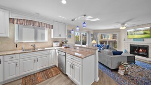 A modern kitchen with white cabinets, a granite countertop, and a built-in island. There is a fireplace with a TV above it, and a living area with a sofa and a rug. - Ruby Villa B - Upper Unit for 6! (Newport Beach)