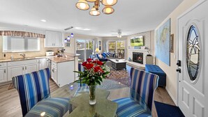 A modern kitchen with white cabinets, a dining area with striped blue chairs, a living room with a fireplace, and a large window with blinds. - Ruby Villa B - Upper Unit for 6! (Newport Beach)
