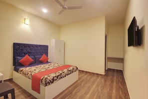 Ein Schlafzimmer mit einer gepolsterten Kopfteile, einem Bett mit rot-gemusterten Bettwaren, einem weißen Kleiderschrank, einem Deckenventilator und einem an der Wand montierten Fernseher.