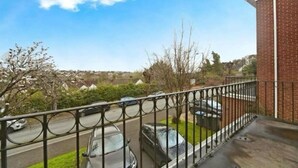 Ein Balkon mit Metallgeländer, der auf eine Straße mit parkenden Autos und ein Backsteingebäude im Hintergrund blickt.