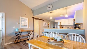 A dining area with a wooden table and chairs, a kitchen in the background, and a clock on the wall. - Highland Shores Haven Fit For 7 (Lake Ozark)
