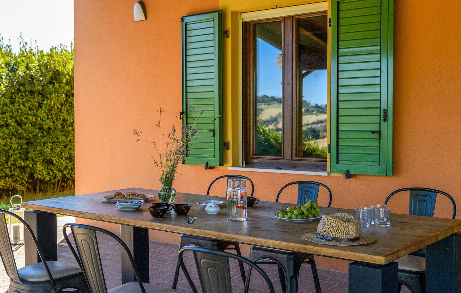 Mesa lista para comer con vista al campo a través de una ventana con contraventanas verdes.