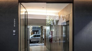 Une entrée moderne avec une porte en verre, une voiture visible à l’extérieur et un arbre en arrière-plan.