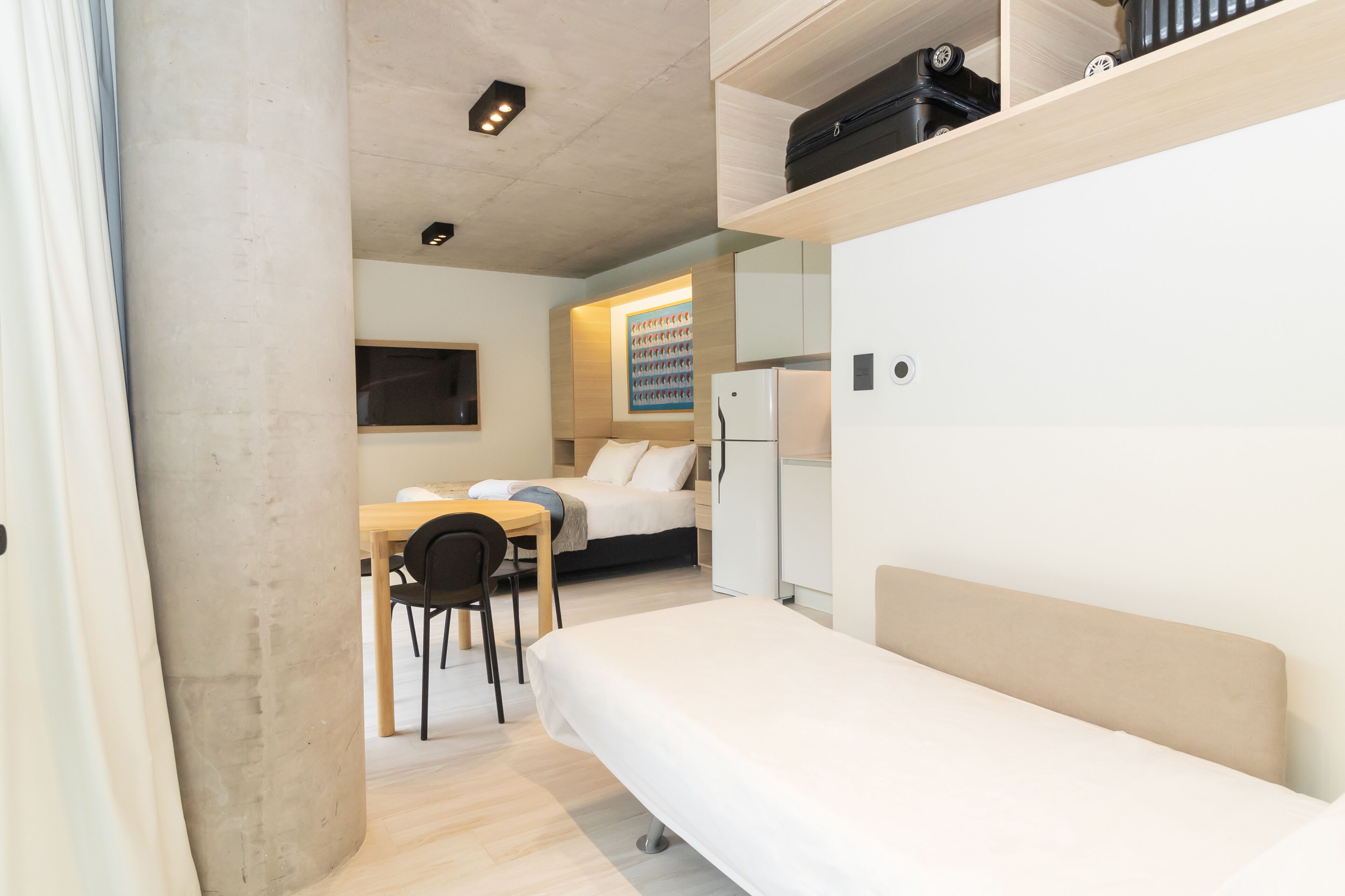 A compact room with a bed, a desk, and a chair. There is a refrigerator and a shelf with items on it.