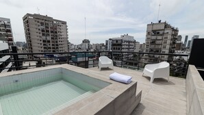 Un espace piscine aménagé sur le toit, avec un jacuzzi, deux chaises en plastique blanches et une vue sur le paysage urbain.