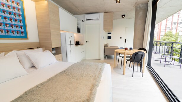 Une chambre d’hôtel moderne équipée d’un lit, d’une petite kitchenette, d’un coin repas avec une table et des chaises, et d’un balcon offrant une vue sur la ville.