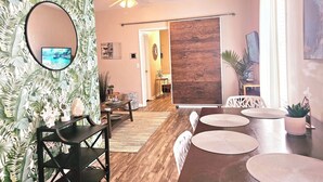 A living room with a wooden floor, a ceiling fan, a dining table with chairs, a TV on a wall, and a large mirror.